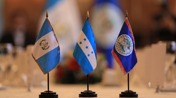 Tres pequeñas banderas de mesa de Guatemala, Honduras y Belice alineadas sobre un mantel blanco.