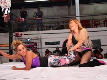 Dos mujeres luchando en un ring de lucha libre, rodeado de cuerdas y con espectadores al fondo