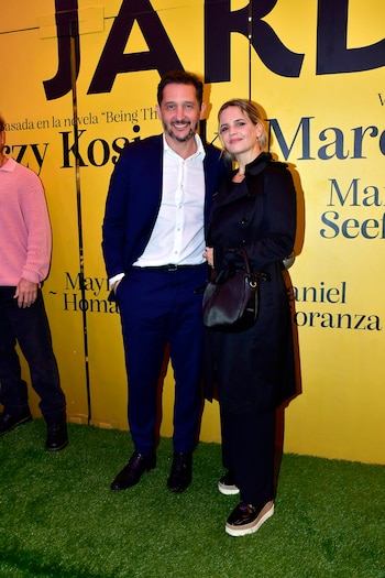 Un hombre con traje azul y una mujer con gabardina negra sonríen, posando en una alfombra de césped artificial verde frente a un fondo amarillo con letras negras grandes en el estreno de la obra