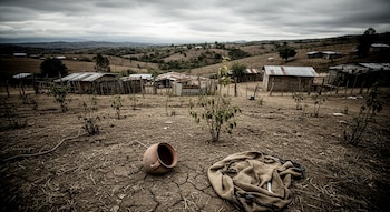 Terreno seco y agrietado con una olla de barro volcada y una manta en primer plano, rodeado de chozas de madera y techos de zinc bajo un cielo nublado.