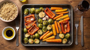 Bandeja con coles de Bruselas, zanahorias, pimientos y ajo asados, junto a un cuenco de quinoa, aceite, cubiertos y una copa de vino tinto.