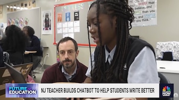 Un hombre con barba y una estudiante afroamericana con trenzas observan una laptop en un aula. Hay otras laptops y carteles educativos en el fondo