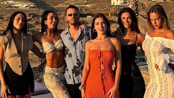 Seis personas, un hombre y cinco mujeres, posan sonrientes al atardecer. Visten ropa de fiesta. Fondo con colinas áridas, casas blancas y palmera