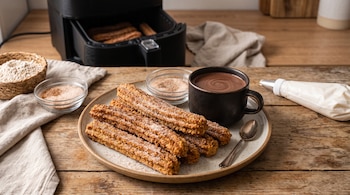 Churros en un plato junto a una taza de chocolate, una freidora de aire abierta y una manga pastelera sobre una mesa de madera.