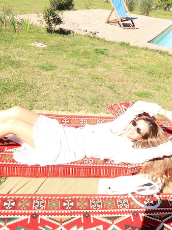 Una mujer con gafas de sol y vestido blanco está acostada sobre una colchoneta roja decorada en el césped junto a una piscina