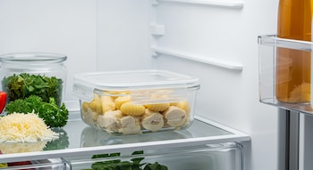 Una heladera blanca exhibe un recipiente de vidrio con ñoquis y pollo a la crema, perejil, queso rallado, un frasco con hierbas y una botella de líquido ámbar.