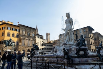 Vista diurna de la Fuente de Neptuno en Florencia con su estatua central de mármol y figuras de bronce, salpicaduras de agua, edificios históricos y personas al fondo