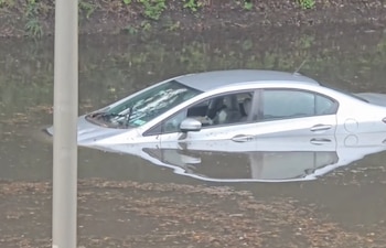 Coche sedán plateado parcialmente sumergido en agua turbia. Se observan las ventanas y el techo del vehículo, con un poste vertical a la izquierda