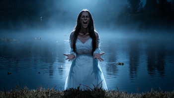 Una mujer con vestido blanco grita con las manos abiertas mientras permanece en el agua rodeada de niebla en un entorno nocturno