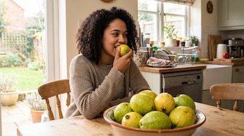 Mujer joven de cabello rizado come una guayaba verde y amarilla en una mesa de madera, con un frutero lleno de guayabas y una cocina al fondo.
