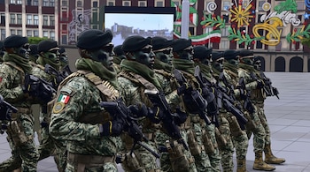 Fila de soldados con uniforme de camuflaje, boinas negras, gafas de sol, maquillaje facial, parches de bandera mexicana y armas. Fondo con edificios.