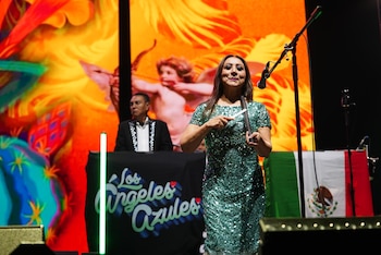 Mulher com vestido de lantejoulas verdes tocando güiro no palco leve. Ao fundo, um homem e uma tela animada, ao lado da bandeira mexicana
