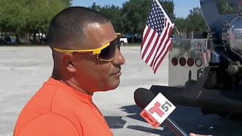 Primer plano lateral de un hombre con camisa naranja y gafas de sol amarillas, hablando a un micrófono de noticias, con una bandera de Estados Unidos detrás