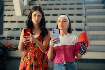 Dos mujeres jóvenes posan, una con un guante de boxeo rojo y la otra sosteniendo un teléfono rojo, frente a una pared estructurada y escaleras