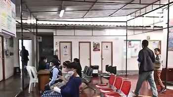 Interior de una posta médica con sala de espera. Varias personas sentadas en sillas rojas y oscuras; otras de pie. Techo metálico y puertas blancas visibles