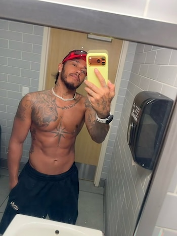 Un hombre sin camiseta con tatuajes se toma una selfie en el espejo de un baño con paredes de azulejos grises, un lavabo blanco y un dispensador negro
