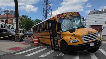 Autobús escolar amarillo inclinado en un paso de cebra. Conos de tráfico y cinta de precaución. Vehículos y una torre de comunicaciones al fondo
