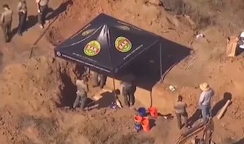 Vista aérea de personas en una fosa excavada junto a una carpa negra y varios cubos en un terreno de tierra