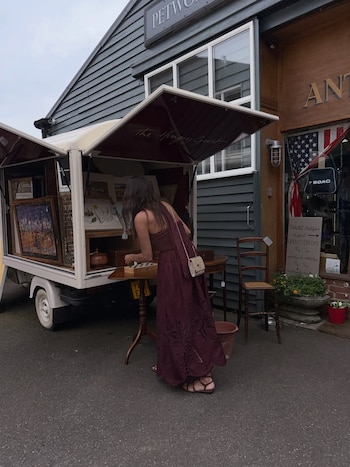 Mujer con vestido borgoña se inclina para ver artículos en camión de venta ambulante y mesa de madera. Edificio gris y tienda de antigüedades en el fondo