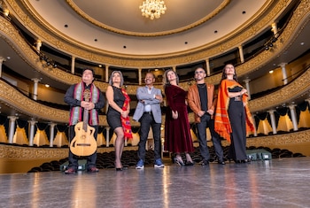 Seis personas vestidas con trajes elegantes posan sobre un escenario teatral de madera ante palcos dorados y una araña de luces