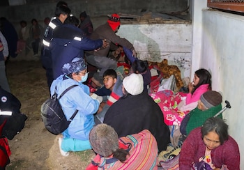 Grupo de personas sentadas en el suelo sobre mantas junto a un muro. Incluye adultos y niños. Un adulto en uniforme azul y mascarilla se inclina hacia ellos