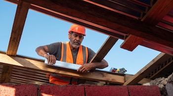 Un obrero con casco naranja y chaleco reflectante manipula una lámina metálica sobre vigas de madera bajo un cielo despejado.