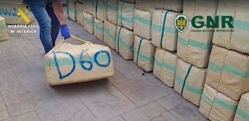 Una persona con guantes levanta un saco de tela marcado con la inscripción D60 frente a varias filas de paquetes apilados