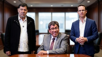 Un hombre con barba de pie a la izquierda, un hombre sonriente con gafas sentado al centro y un hombre con traje de pie a la derecha, en una sala de juntas.