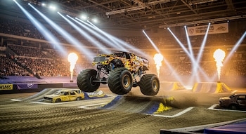 Un monster truck con diseño colorido y ruedas gigantes está en pleno salto sobre un coche destrozado en el centro de una arena, con focos brillantes e iluminación escénica, y tribunas llenas de espectadores al fondo. Fuego sale de dispositivos en el suelo.