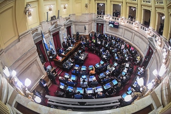 Vista aérea del hemiciclo del Senado de la Nación Argentina, con senadores sentados en escaños con pantallas, en una sala con alfombra roja y arquitectura clásica