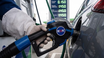 Mano enguantada sostiene pistola de surtidor de combustible con logo RECOPE, llenando tanque de auto. Fondo desenfocado con display digital mostrando precios en descenso.