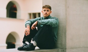 Retrato de Nelio Biedermann, un hombre joven con bigote, sentado en un banco de piedra con una sudadera verde y pantalones oscuros, mirando al frente