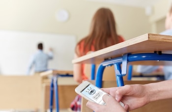 Primer plano de manos sosteniendo un teléfono móvil con una aplicación de chatbot abierta. Al fondo, estudiantes y un profesor en un aula con pupitres y una pizarra