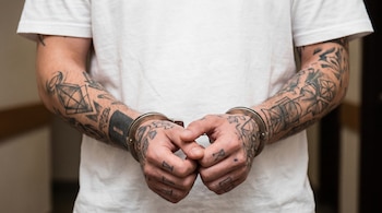 Manos de persona con tatuajes, esposadas con grilletes, brazos tatuados y camiseta blanca.