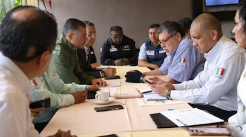 Nueve personas, hombres y una mujer, sentadas alrededor de una mesa beige con documentos, cuadernos, bolígrafos, teléfonos móviles y una taza.
