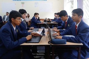 Varios estudiantes jóvenes, vistiendo uniformes de chaqueta azul, sentados en pupitres en un aula, escribiendo en laptops. Adultos en el fondo