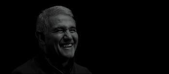 Retrato en blanco y negro de un hombre de mediana edad sonriendo ampliamente, con cabello gris y arrugas faciales, contra un fondo negro liso