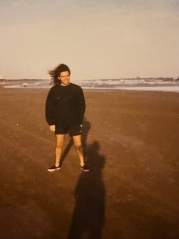 Hombre joven de cabello largo y oscuro, con sudadera y pantalones cortos, de pie en una playa de arena con el mar al fondo y el cielo claro