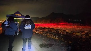 Funcionarios policiales con chaquetas operativas caminan de noche junto a un terreno iluminado con luces rojas de emergencia