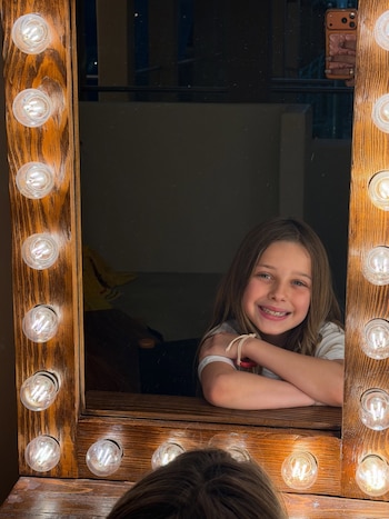 Reflejo de una niña sonriendo en un espejo de tocador con marco de madera y bombillas encendidas alrededor