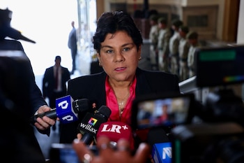 Norma Yarrow frente a micrófonos de prensa y un teléfono grabando, con varias personas y militares borrosos en el fondo