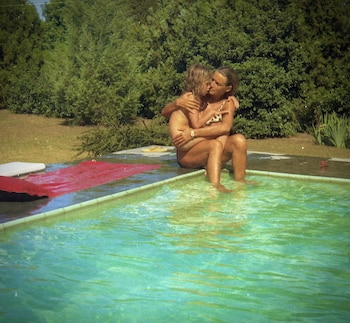 Dos mujeres se abrazan en el borde de una piscina, con los pies sumergidos en el agua. Al fondo, una pared de arbustos verdes bajo un cielo claro