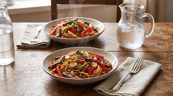 Dos platos de fideos con tiras de carne, pimientos rojos y ají. Una botella de agua y una jarra de agua con hielo, y tenedores con servilletas en mesa de madera.