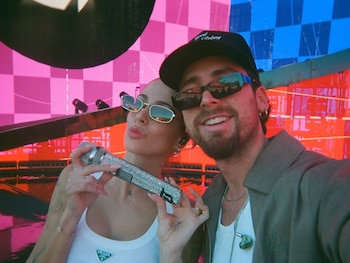 Primer plano de Tini Stoessel con gafas y un micrófono brillante, junto a un hombre barbudo con gorra. Fondo de escenario con pantallas rosas y azules