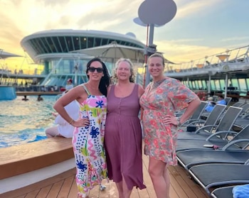 Tres mujeres sonríen de pie en la cubierta de un crucero junto a una piscina y tumbonas al atardecer
