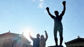 Siluetas de dos personas levantando los brazos junto a la estatua de Rocky, con un cielo azul brillante y un edificio clásico de fondo