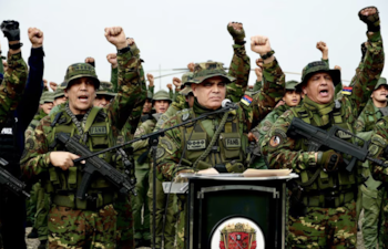 Militares venezolanos en uniforme de camuflaje. Varios alzan los puños. Dos oficiales al frente, uno en un podio con micrófono y otro sosteniendo un arma