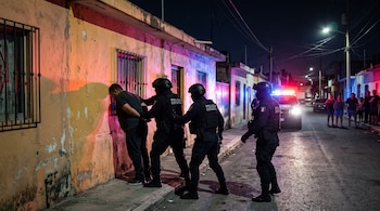 Tres policías estatales con equipo táctico sujetan a un hombre contra la fachada de una casa durante la noche mientras vecinos observan.