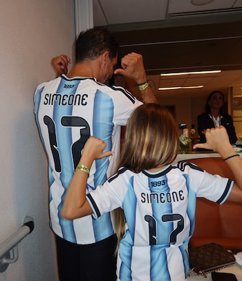Hombre adulto y niña de espaldas con camisetas de fútbol celestes y blancas que dicen 'Simeone' y '17'. Ambos hacen gesto de pulgar hacia abajo