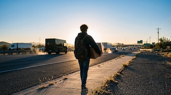 Vista posterior de una persona que camina por la acera de una carretera llevando una mochila al hombro y una caja de cartón.